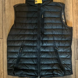 Svart dunväst från Parajumpers - Snygg svart dunväst från Parajumpers med quiltad design och dragkedja framtill. Västen har två snedställda fickor med dragkedja och en hög krage. Insidan är ljusgrå och har autenticitetsmärke. Perfekt för lager-på-lager-stil.