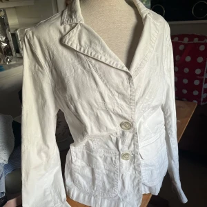 Vit jacka i bomull - Snygg vit kavaj med två knappar framtill och stora fickor. Jackan har ett diskret strukturerat mönster i tyget som ger en cool touch. Klassisk krage och långärmad modell. Går som en M/L i storleken