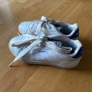 Vita Reebok sneakers med blå detalj - Klassiska vita Reebok sneakers med blå häl och logga. Skorna har snörning, perforerade sidor för extra ventilation och en platt sula. Perfekta för dig som gillar en clean och sportig stil. Materialet är syntet och skinnliknande yta.