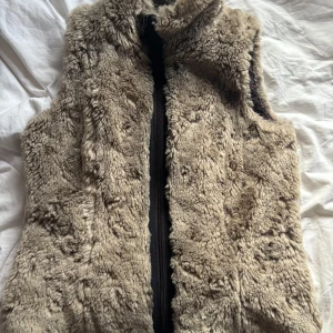 Beige pälsväst från Esprit - Säljer en fluffig beige pälsväst från Esprit med dragkedja framtill. Västen har ett mjukt och mysigt foder och insidan är fodrad med paisleymönstrat tyg i mörka toner. Perfekt att slänga över en tröja för extra värme och stil.