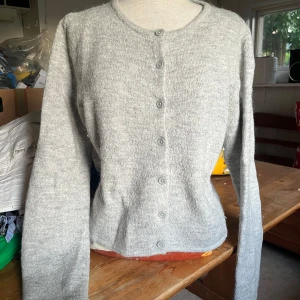 Ljusgrå stickad kofta med knappar - Mysig ljusgrå stickad kofta med rund hals och knäppning framtill. Koftan har långa ärmar och en enkel, klassisk design som passar perfekt till lager-på-lager. Materialet känns mjukt och värmande, perfekt för kyligare dagar. Ordinarie 400kr
