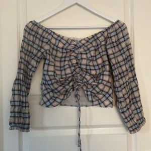 Rutig offshoulder topp från FB Sister - Superfin rutig topp från FB Sister i blått, beige och svart. Offshoulder-modell med långa ärmar och snörning framtill som ger en snygg rynkad effekt. Croppad passform och lätt, luftig känsla – perfekt för en trendig look.