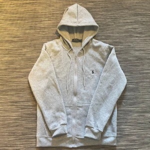 Grå hoodie från Polo Ralph Lauren - Snygg ljusgrå hoodie från Polo Ralph Lauren med dragkedja och huva. Klassisk broderad logga på bröstet och praktiska fickor framtill. Tillverkad i mjuk bomullsmix, perfekt för chill dagar eller när du vill ha en clean look.