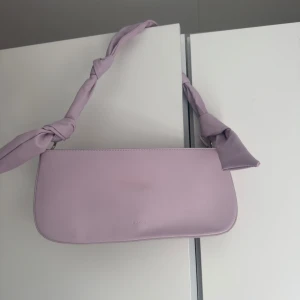 Ljuslila axelväska från NA-KD - Supersöt ljuslila axelväska från NA-KD med stilren, avlång form och knutdetaljer på axelbandet. Väskan har dragkedja upptill och ett innerfack för småsaker. Perfekt att slänga över axeln för en trendig look. Väskan är helt ny och inte används en enda gång. Pris kan diskuteras