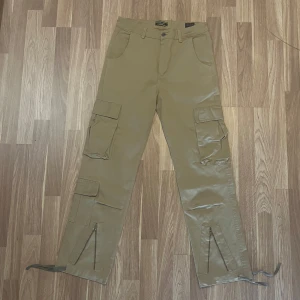 Beiga cargobyxor - Snygga beiga cargyxor med flera stora fickor på sidorna och baktill. Byxorna har raka ben med snörning och dragkedja vid bensluten för extra stil. Byxorna har nån fläck på sig, därav ett lägre pris!
