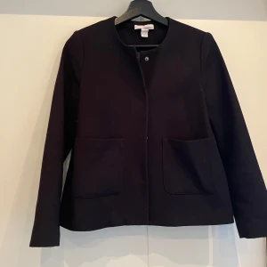 Svart kavaj från H&M, stl 36 - Stilren svart kavaj med struktur från H&M i storlek 36. Rak modell med rund halsringning, två stora fickor framtill och en snygg slits bak. Perfekt för dig som gillar minimalistisk look och vill ha en klassisk jacka i garderoben.