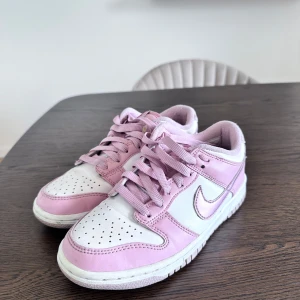 Nike Dunk sneakers i rosa och vitt - Snygga Nike Dunk sneakers i en fräsch kombination av ljusrosa och vitt. Skorna har klassisk låg siluett, perforerad tå och tydlig Nike-logga på sidan. Perfekta för dig som vill sticka ut med en färgglad och trendig look.