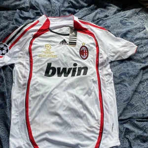 AC Milan Champions League 2007 tröja - AC Milan bortatröja från Champions League-finalen 2007 i Aten. Vit med röda och svarta detaljer, broderat klubbmärke och Champions League-logga på ärmen. Adidas stripes på axlarna och Bwin som sponsor. Tillverkad i lätt och ventilerande polyester. Storleken är L vilket passar dom som är 175-180 cm lång. Helt nyskick lappar finns kvar.  Vid minsta fundering eller om du vill ha mer bilder på tröjan hör av dig.