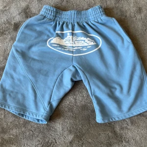 Blåa mjukisshorts med tryck från Corteiz - Säljer ett par ljusblå mjukisshorts från Corteiz med resår i midjan och ett stort vitt tryck av en båt på framsidan. Shortsen har en bakficka och är tillverkade i mjukt bomullsmaterial. Perfekta för chill dagar eller träning.