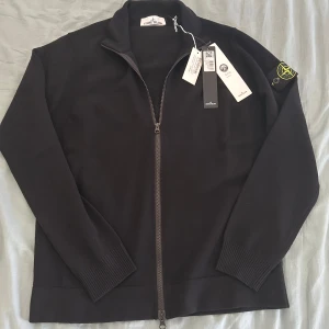 Stickad Stone Island tröja - Svart cardigan tröja från Stone Island med hel dragkedja och klassisk logotyp på ärmen. Tröjan är stickad i mjukt material och har ribbade muddar vid ärmslut och nederkant. Perfekt för dig som gillar stilren och clean design.