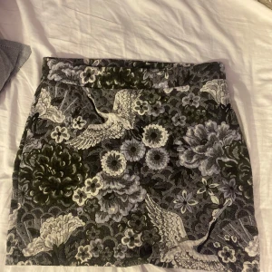 Blommig kortkjol från object  - Supersnygg kortkjol  med unikt blommönster och fåglar i svart, grått och grönt. Kjolen har en rak passform och är perfekt för dig som vill sticka ut med en cool och detaljrik look. Tjockare tyg med vävd känsla.