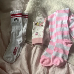 3 stycken Sanrio och randiga strumpor - Tre par söta strumpor: ett par vita Hello Kitty-strumpor med röda ränder, ett par vita och rosa prickiga Sanrio-strumpor från Miniso, samt ett par knähöga randiga strumpor i rosa och vitt. De rosa vita randiga knähöga strumporna är köpta secondhand men aldrig använda, de andra är helt nya! 20kr styck och 50kr för alla.🤍
