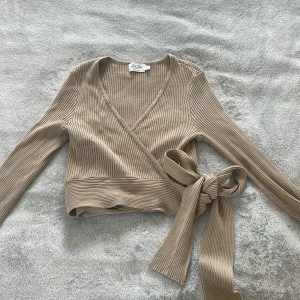 Beige omlott kofta från NA-KD X Anika Teller - Superfin ribbad beige omlott kofta från NA-KD x Anika Teller. Tröjan har långa ärmar, v-ringning och knytband i midjan (som var svårt att knyta snyggt på bild😅). Storlek S.