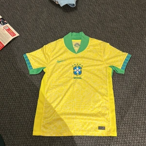 Brasilien Nike fotbollströja gul M - Säljer en officiell Brasilien fotbollströja från Nike i storlek M. Tröjan är gul med gröna detaljer vid krage och ärmslut, samt blå Nike-logga och Brasilien-emblem på bröstet. Tillverkad i Dri-FIT-material som andas. Snyggt grafiskt mönster över hela tröjan.