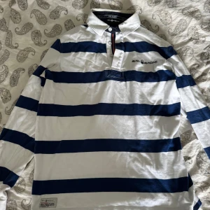 Randig pikétröja från Sail Racing - Snygg långärmad pikétröja från Sail Racing med breda marinblå och vita ränder. Klassisk krage, knäppning vid halsen och broderad logga på bröstet. Perfekt för en sportig och avslappnad stil.