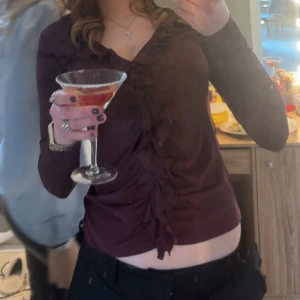 Vinröd topp med volangdetaljer - Säljer en vinröd långärmad topp med snygga volangdetaljer längs v-ringningen och mitt fram. Toppen har en figurnära passform och är perfekt för dig som vill ha något stilrent men ändå med en twist. Superfin färg som passar till mycket!