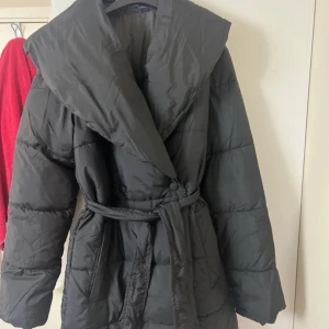 Svart pufferjacka med bälte - Säljer en svart pufferjacka med bred krage och bälte i midjan. Jackan är quiltad och har en oversized känsla, perfekt för kalla dagar. Långärmad och tillverkad i syntetmaterial. Enkel och stilren design som håller dig varm hela vintern. Inte använd en ända gång men den har inte prislapp. 