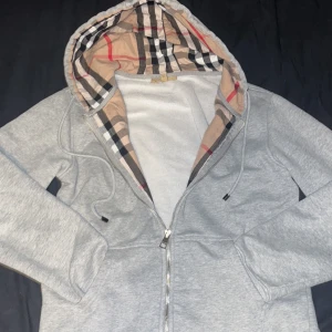 Grå Burberry hoodie med rutigt foder - Säljer en grå hoodie från Burberry med dragkedja och klassiskt rutigt foder i huvan. Tröjan har snörning vid huvan och broderad logga på bröstet och även de rutiga mönstert på insidan. Mycket fint skick endast använd fåtal gånger! Storlek L men passar lite bättre till M och S