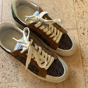 Bruna sneakers från Zadig & Voltaire - Snygga sneakers från Zadig & Voltaire med bruna mockadetaljer och mönstrade paneler i svart och brunt. Skorna har vita snören, vit sula och en cool retrovibe. Perfekta för dig som gillar streetstyle och vill sticka ut med ett unikt mönster.