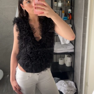 Svart volangväst med v-ringning - Säljer en svart body med stora volanger och djup v-ringning. Bodyn är ärmlös och har en fluffig, festlig look som sticker ut. Perfekt för dig som vill ha något unikt och trendigt i garderoben.