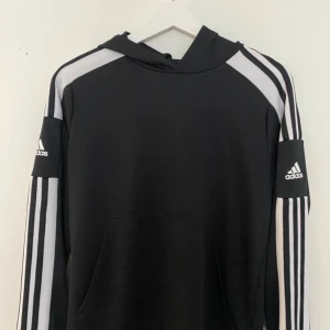 Svart hoodie från Adidas med vita ränder - Säljer en svart hoodie från Adidas med klassiska vita ränder längs ärmarna och Adidas-logga på båda ärmarna. Tröjan har huva och en stor magficka framtill. Perfekt för en sportig och avslappnad stil.Aldrig använd