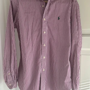 Röd- och vitrandig skjorta från Ralph Lauren - Säljer en klassisk röd- och vitrandig skjorta från Ralph Lauren med knappar framtill och broderad logga på bröstet. Skjortan har lång ärm och krage, perfekt för en stilren look. Skjortan är i bra skick.