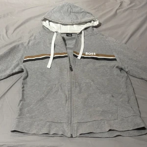 Grå hoodie med dragkedja från BOSS - Grå hoodie från BOSS med dragkedja framtill och huva med vita snören. Brunt, vitt och svart band över bröstet samt BOSS-logga. Perfekt för en avslappnad stil. Köpt för 1200kr