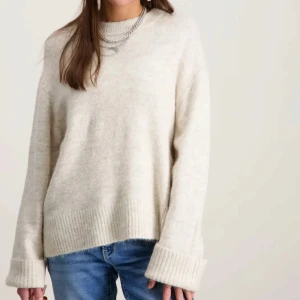 Beige stickad tröja med rund hals - Mysig beige stickad tröja med rund hals och breda ribbade muddar vid ärmslut och nederkant. Tröjan har en avslappnad passform och långa ärmar, perfekt för lager-på-lager-stil. Storlek S