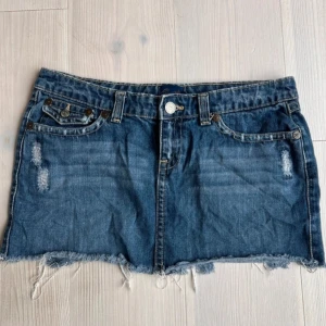 Jeanskjol med fransig kant - Säljer en klassisk blå jeanskjol med fransig, rå nederkant och slitna detaljer framtill. Kjolen har fem fickor och knappstängning. Perfekt för dig som gillar en avslappnad och trendig stil.