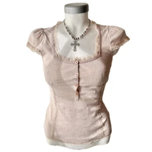 Beige topp med spets och knappar - Söt beige topp med korta puffärmar, spetsdetaljer runt halsringningen och dekorativa knappar framtill. Kvadratisk urringning och figurnära passform. Perfekt för en romantisk och feminin stil.