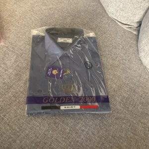 Blå skjorta från Golden 2000 - Säljer en klassisk blå skjorta från Golden 2000 med krage och knappar framtill. Skjortan har ett broderat kronmotiv på bröstet och är perfekt för dig som gillar stilrena plagg. Levereras i originalförpackning.