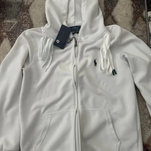 Vit hoodie från Polo Ralph Lauren - Säljer en klassisk vit hoodie från Polo Ralph Lauren med dragkedja och huva. Tröjan har två fickor framtill och den ikoniska broderade loggan på bröstet. Perfekt för en clean och stilren look. Pris kan såklart diskuteras, kommer säkert acceptera era prisförslag sålänge det är inte överdrivet lite eftersom behöver pengarna skriv gärna