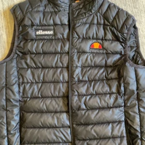 Svart dunväst från Ellesse - Snygg svart dunväst från Ellesse med quiltad design och dragkedja framtill. Västen har Ellesse-logga på bröstet och passar perfekt som ett extra lager under kyliga dagar.