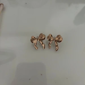 Guldiga rosettörhängen - Snygga örhängen i form av små rosetter i guldig färg. Perfekta för att ge en söt och trendig touch till din look. Stiftmodell som sitter bekvämt på örat.
