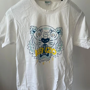Vit t-shirt med tigertryck från Kenzo - Vit t-shirt från Kenzo med ikoniskt tigertryck och Kenzo Paris-logga i gult och blått framtill. Klassisk passform och rund halsringning. Perfekt för dig som vill ha en statement-tee med cool design. Köpte för: 900kr säljer för 450kr