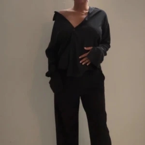 Svart oversized långärmad blus med kostym byxa  - Säljer en svart oversized blus med v-ringning och långa ärmar. Blusen har en lös passform och bred krage som kan bäras offshoulder. Perfekt för en avslappnad men stilren look. Armarna är snyggt slittade, kan köpas separat eller ihop med kostym byxorna för hela looken, lapp kvar på tröjan, byxor aldrig använda heller 