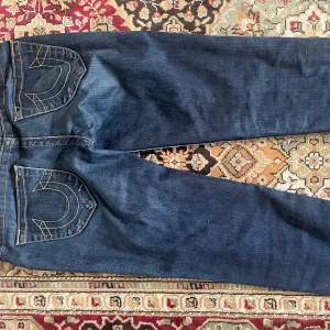Mörkblå jeans från True Religion - Säljer ett par klassiska mörkblå jeans från True Religion med deras ikoniska kontrastsömmar och stora bakfickor med broderad logga. Jeansen har raka ben och normal passform. Perfekta för dig som gillar streetwear och snygga detaljer.