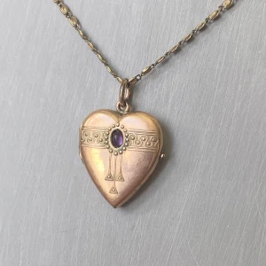 Hjärtformat medaljonghalsband i guld med lila sten - Unikt hjärtformat medaljonghalsband i guldfärgad metall med dekorativa mönster och en lila sten i mitten. Medaljongen går att öppna och har plats för ett foto eller minne. Kedjan är tunn och elegant.