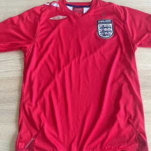 England fotbollströja röd Umbro - Säljer en röd England fotbollströja från Umbro med broderat landslagsmärke och guldstjärna på bröstet. Tröjan har korta ärmar och klassisk v-ringad krage. Den är äkta!