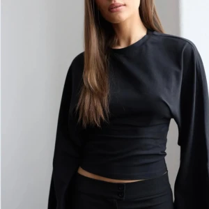 Svart långärmad topp med vida ärmar -  Är slutsåld just nu på Gina Säljer en svart långärmad topp med rund hals och extra vida ärmar. Modellen är figurnära över kroppen och har en croppad passform. Perfekt för dig som vill ha en stilren men trendig look.