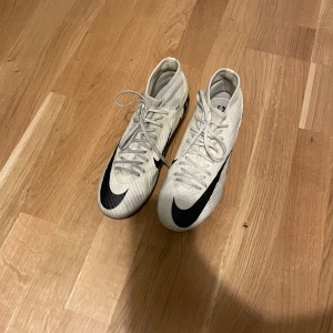 Nike fotbollsskor med snörning - Säljer ett par vita Nike fotbollsskor med svart swoosh och textdetaljer. Skorna har snörning och högre skaft för att det inte ska komma in smuts och andra saker. Perfekta för dig som vill ha stilrena och lätta skor på planen.