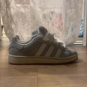 Ljus blå Adidas Campus sneakers - Säljer ett par ljus blå Adidas Campus sneakers med vita ränder och vit sula. Klassisk modell med snörning och låg profil, perfekt för en avslappnad stil. Ikoniska tre ränder på sidan och Campus-logga i silver. Priset kan diskuteras 