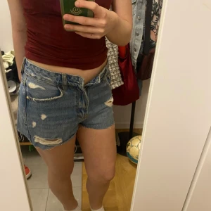 Blå jeansshorts med slitningar - Säljer ett par blå jeansshorts med råa kanter och slitna detaljer. Klassisk femficksmodell med knapp och dragkedja framtill. Shortsen har låg midja och en avslappnad passform. Perfekta för sommaren!