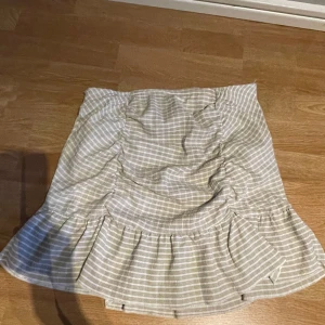 Beige och vit rutig kjol från Gina Tricot - Supersöt kortkjol från Gina Tricot i beige och vitt rutigt mönster. Kjolen har volang nertill och rynkade detaljer framtill som ger en snygg form.