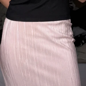 Ljusrosa plisserad kjol - Säljer en ljusrosa plisserad kjol med smala vertikala ränder. Kjolen har en rak passform och är midilång, perfekt för en trendig och stilren look. Materialet är glansigt och ger ett elegant intryck. Det är dubbelt material, där de undre är satin. Står storlek XS, 36.