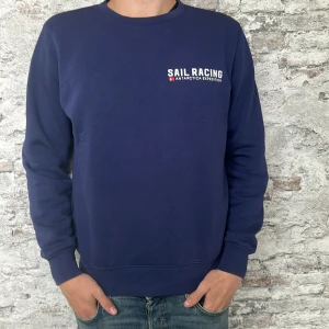 Blå sweatshirt från Sail Racing - Mörkblå sweatshirt från Sail Racing med tryck på bröstet och stor grafisk detalj på ryggen. Rund halsringning och långa ärmar. Perfekt för en sportig och avslappnad stil. |Det är bara och skriva vid intresse|