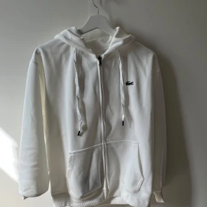 Vit hoodie med dragkedja från Lacoste - Säljer en klassisk vit hoodie från Lacoste med dragkedja framtill och huva med snören. Tröjan har två stora fickor fram och den ikoniska krokodilloggan på bröstet. Perfekt för en avslappnad och stilren look.