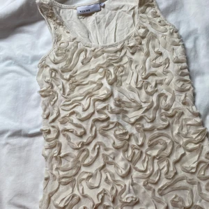 Beige ärmlös klänning med volang från KappAhl - Säljer en beige ärmlös klänning från KappAhl med unikt volangmönster över hela framsidan. Klänningen har rund halsringning. Står storlek xs men skulle säga passar mer S oxh M ifall man vill ha det lite tajt 