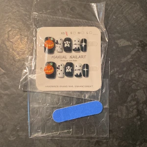 Halloween-inspirerade lösnaglar med nagelfil - Säljer ett set med svarta och vita lösnaglar med Halloweenmotiv som pumpa, spöke och katt. Paketet innehåller även en liten blå nagelfil och självhäftande nageltejp för enkel applicering.