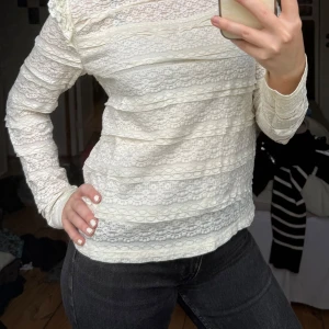 Vit spetsblus med volangdetaljer - Säljer en söt vit långärmad spetsblus med volangdetaljer på axlarna. Blusen har ett romantiskt mönster och är perfekt att styla med jeans eller kjol. Materialet är tunt och genombrutet vilket ger en luftig känsla. Tröjan är från Daisy Grace. 